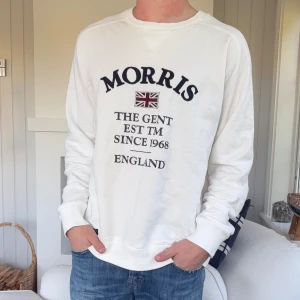 Vit sweatshirt från Morris Gents - Vit sweatshirt från Morris Gents med tryck framtill och broderad Union Jack-detalj. Tröjan har rund hals, långa ärmar och ribbade muddar. Tillverkad i mjuk bomull för en skön och avslappnad stil.