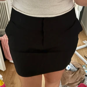 Svart kortkjol med fickor - Snygg svart kortkjol med stilren design och diskreta fickor framtill. Kjolen har en tight passform som framhäver figuren och är tillverkad i ett mjukt material som känns bekvämt mot huden. Perfekt för dig som gillar minimalistisk stil och vill ha en enkel men trendig kjol.