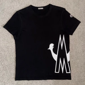 Svart Moncler t-shirt med stor logga - Svart t-shirt från Moncler i bomull med stor vit logga och tupp-tryck på framsidan. Klassisk rund halsringning och korta ärmar. Moncler-märke på ärmen och autenticitetsmärke på insidan. Snygg och stilren design som sticker ut. Storleken är S men sitter som XS. Nypris: 4000kr💯✅