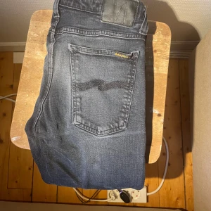 Svarta jeans från Nudie Jeans - Säljer ett par svarta jeans från Nudie Jeans med klassisk femficksdesign och snygga detaljer på bakfickan. Jeansen har en mörk tvätt och är tillverkade i mjukt denimtyg som sitter skönt. Perfekt för dig som gillar stilrena och tidlösa jeans. En av rämmarna där man har bältet är sönder där bak men går att sy ihop lätt!