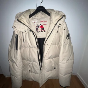 Beige pufferjacka Moose Knuckles XL - Säljer en beige pufferjacka från Moose Knuckles i storlek XL. Jackan har stor huva fodrad med mjuk fleece, svarta dragkedjor och logga på ärmen. Perfekt för kalla dagar med sin dunfyllda insida och rejäla fickor framtill.