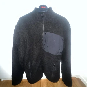 Svart teddyjacka med dragkedjor - Svart teddyjacka med hög krage och hel dragkedja framtill. Jackan har en stor bröstficka i nylon med dragkedja samt två sidofickor med dragkedjor. Mjuk och fluffig känsla, perfekt för kyliga dagar.