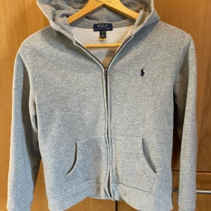 Grå hoodie från Polo Ralph Lauren - Klassisk grå hoodie från Polo Ralph Lauren med dragkedja, huva och känguruficka. Broderad marinblå logga på bröstet. Mjuk insida men är lite uttöjd i ärmarna men är inget som syns. perfekt för chill dagar. Snygg och enkel att matcha med allt. Sitter som xs i herrstorlek! Pris kan diskuteras.