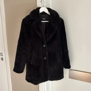 Svart pälsjacka från Pull&Bear - Mysig svart pälsjacka från Pull&Bear med bred krage och två knappar framtill. Jackan har två sidofickor och den ena av dem är trasig invändigt. 