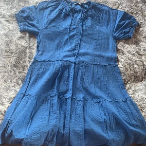 Blå klänning med volanger - Supersöt blå klänning med volanger och struktur i tyget. Klänningen har korta puffärmar med resår, knytning i halsen och flera lager för en flowy känsla. Perfekt för dig som gillar en avslappnad men ändå söt stil.Är använd en gång och har ett litet märke på en arm, vilket inte märks när man har på sig klänningen.Är en luftig klänning, säljer för den är lite tajt över bysten på mig. 
