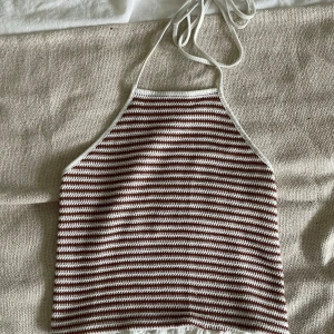 Randig croppad linnetopp med knyt - Säljer en croppad linnetopp med smala axelband som knyts i nacken. Toppen har ett randigt mönster i vitt och brunt, och är tillverkad i ett stickat material. Baksidan har en smockad panel med volangkant för extra stretch och snygg passform.