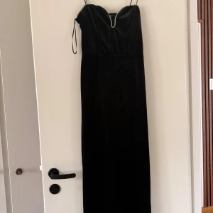 Svart långklänning med smala axelband - Elegant svart långklänning med smala axelband och en snygg v-ringning dekorerad med en glittrig detalj. Klänningen har en figurnära passform och är tillverkad i ett mjukt sammetstyg som ger en lyxig känsla.