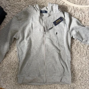 Grå hoodie från Polo Ralph Lauren - Snygg grå hoodie från Polo Ralph Lauren med dragkedja framtill och klassisk huva. Tröjan har två fickor och den ikoniska broderade loggan på bröstet. Perfekt för chill dagar och enkel att matcha med jeans eller joggers.