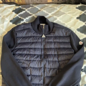 Moncler cardigan - Snygg svart cardigan från Moncler med quiltad framsida och ribbad krage. Västen har dragkedja framtill, två fickor med dragkedja och Moncler-logga på axeln. Perfekt för lager-på-lager och stilren look. Den är storlek XL men kan passa som L. Jag har inte använt eftersom att den passade inte mig.