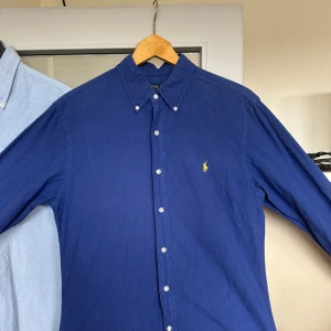 Marinblå skjorta från Polo Ralph Lauren - Klassisk marinblå skjorta från Polo Ralph Lauren med button-down krage och broderad gul logga på bröstet. Skjortan har långa ärmar och vita knappar framtill. Perfekt för en stilren och tidlös look. Även väldigt försiktigt använd.