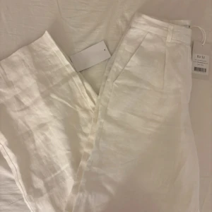 Vita linnebyxor från NA-KD, storlek 32 - Snygga vita linnebyxor från NA-KD med hög midja och vida ben. Byxorna har pressveck framtill, hällor för bälte och två bakfickor med knapp. Perfekta för en fräsch och stilren look under varmare dagar.