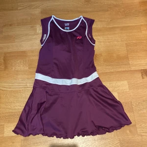 Yonex - Vinröd sportklänning - Badminton  - Sportig vinröd klänning från Yonex med vita detaljer och markerad midja. Ärmlös modell med rund halsringning och inbyggd bh. Klänningen har en lätt utsvängd kjol och knytband i ryggen, perfekt för dig som gillar att sticka ut på tennis eller badmintonbanan.