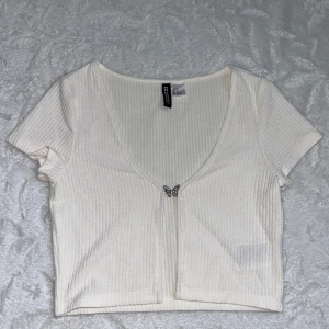 Vit croppad topp från H&M - Säljer en vit croppad topp från H&M. Toppen har djup v-ringning och stängs framtill med en söt liten fjärilsdetalj i metall. Aldrig använd 🥰