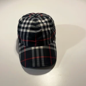 Rutig keps från Burberry - Snygg keps från Burberry med klassiskt rutigt mönster i svart, vitt och rött. Kepsen har böjd skärm, justerbar rem baktill med metallspänne och är tillverkad i bomull. Perfekt accessoar för en trendig streetstyle-look.