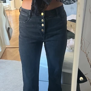 Svarta bootcut jeans med knappar - Snygga svarta jeans med hög midja och bootcut-ben. Byxorna har fem guldiga knappar framtill och klassiska fickor både fram och bak. Perfekta för dig som gillar en stilren look med lite edge. Använd 1 gång och är som nya.