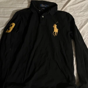 Svart långärmad piké från Polo Ralph Lauren - Svart långärmad pikétröja från Polo Ralph Lauren med stor guld broderad logga på bröstet och siffran 3 på ärmen. Klassisk krage med knappar och tydlig POLO-tryck i gult på ryggen. Perfekt för en sportig och stilren look.