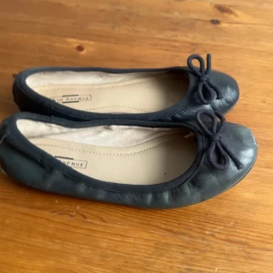 Svarta ballerinaskor med rosett .36 - Snygga svarta ballerinaskor från 5th Avenue i mjukt skinn. Skorna har rund tå och en söt rosettdetalj framtill. Perfekta att slänga på sig till jeans eller klänning för en enkel och stilren look.