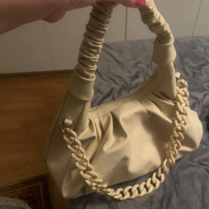 Beige axelväska med kedjedetalj - Trendig beige axelväska i syntetmaterial med snygga rynkade detaljer på handtaget och en chunky kedja i plast. Väskan har en mjuk, slouchy form och stängs med dragkedja. Perfekt för dig som gillar stilrena accessoarer med en twist.