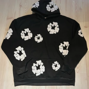 Svart hoodie från denim tears - Svart hoodie från Denim Tears med stora vita blommönster över hela tröjan. Klassisk huva och känguruficka framtill. Mjuk och skön bomullskvalitet, perfekt för en chill och trendig look.                                                                                Priset kan diskuteras 