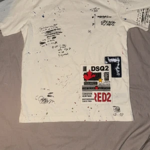 Vit DSQUARED2 t-shirt - Vit t-shirt från DSQUARED2 med coola tryck, handskrivna texter och färgstänk över hela plagget. Stor patch med logga och röd lönnlöv på framsidan. Klassisk passform och korta ärmar. Tillverkad i mjuk bomull för skön känsla.