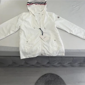 Vit vindjacka från Moncler med huva - Snygg vit vindjacka från Moncler med huva som har röd och marinblå rand. Jackan har dragkedja framtill, snörning vid huvan och två fickor med knapp. Moncler-logga på ärmen ger extra stilpoäng. Perfekt för dig som gillar clean och sportig look.