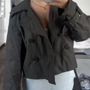 Mörkgrön croppad trenchcoat från H&M - Snygg croppad trenchcoat från H&M i mörkgrön färg. Jackan har dubbelknäppning, bred krage och bälte med spänne vid ärmsluten. 