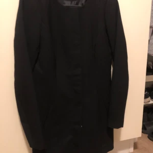 Svart lång kappa med knappar - Stilren svart kappa med lång passform och diskreta fickor på sidorna. Kappan har raka linjer, dold knäppning med tryckknappar och är helfodrad. Perfekt för dig som gillar en clean och klassisk look.