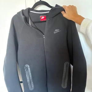 Svart Nike hoodie med dragkedja XS - Svart hoodie från Nike i storlek XS med hel dragkedja framtill, två fickor med dragkedja och klassisk Nike-logga på bröstet. Tröjan har huva och är tillverkad i mjuk bomullsblandning, perfekt för chill eller träning. Men har en defekt bredvid fickan 