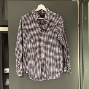 Rutig skjorta från Ralph Lauren - Snygg rutig skjorta från Ralph Lauren i blått och vitt med klassisk button-down krage och orange broderad logga på bröstet. Skjortan har långa ärmar, knappar framtill och är tillverkad i mjuk bomull. Perfekt för en stilren och avslappnad look.