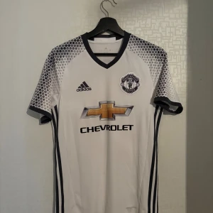Manchester United fotbollströja Paul Pogba  - Manchester United bortatröja 2016/17 – Pogba #6 Original Adidas-tröja (produktkod: AI6690). Premier League-tryck på ryggen (POGBA 6).  Sponsor: Chevrolet. Storlek: XS (passar även S). Skick: Mycket bra (sömmarna lossnar längst ned). Snygg och sällsynt bortatröja från Paul Pogbas första säsong tillbaka i Manchester United, efter att han köptes för dåvarande världsrekord summa. Perfekt för samlare eller United-fans! 