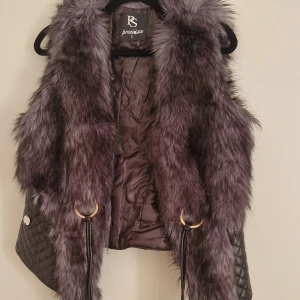 Svart pälsväst med quiltade detaljer - Snygg svart pälsväst från Preziosa med quiltade skinnpartier nertill och coola guldfärgade ringar med långa snören framtill. Västen har en fluffig känsla och mixar fuskpäls med glansigt material för en riktigt trendig look.