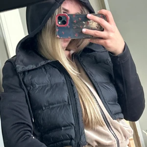 Svart croppad pufferjacka med huva - Svart croppad pufferjacka med huva och dragkedja framtill. Jackan har quiltad front, släta ärmar och snörning i midjan. Perfekt för dig som gillar en sportig och trendig look. Materialet är syntet och polyester.