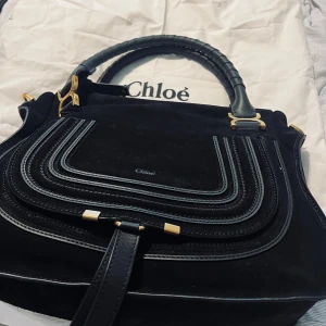 Svart axelväska i mocka - Axelväska i lyxig mocka med detaljer i skinn och guldfärgade metalldelar. Väskan har ett snyggt handtag med lindat skinn och dekorativa sömmar framtill. Perfekt för dig som vill ha en stilren och exklusiv väska.