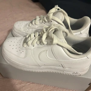 Nike Air Force 1 vita sneakers - Klassiska Nike Air Force 1 sneakers i helvitt skinn med perforerad tå, rund tå och platt sula. Skorna har snörning och den ikoniska swoosh-loggan på sidorna. Perfekta för dig som gillar en clean och tidlös look.