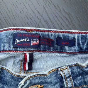 Slim jeans från U.S. Polo Assn. - Snygga blå jeans från U.S. Polo Assn. i slim fit-modell med klassisk femficksdesign och broderad logga på ena benet. Jeansen har kontrastsömmar och detaljer i rött och vitt vid midjan. Perfekt för dig som gillar en sportig och stilren look.