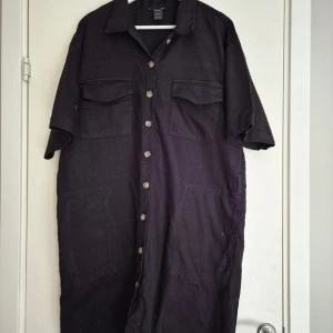 Svart skjortklänning från Lindex - Svart skjortklänning från Lindex i 100% bomull. Modellen är rak och loose fit med korta ärmar, knappar hela vägen framtill, två bröstfickor med lock och två stora fickor framtill. Skärp saknas.