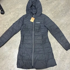 Mörkgrå dunjacka från Seaside - Snygg mörkgrå dunjacka från Seaside med huva och lång passform. Jackan har quiltad design, dragkedja framtill och långa ärmar. Perfekt för kalla dagar och enkel att matcha med din stil. Märket syns diskret på bröstet.