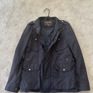 Mörkblå parkajacka från Woolrich - Säljer en svart parkajacka från Woolrich med flera stora fickor framtill och snygga silverfärgade knappar. Jackan har dragkedja och tryckknappar, samt en klassisk krage. Perfekt för dig som vill ha en stilren och funktionell jacka.