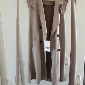 Beige trenchcoat från Zara med bälte - Stilren beige trenchcoat från Zara med klassisk krage, knappar framtill och tillhörande bälte i midjan. Jackan har fickor på sidorna och är tillverkad i ett mjukt material som ger en snygg och avslappnad look. Helt nya med lappen kvar 
