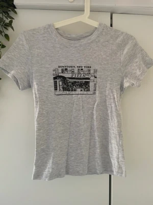 Grå t-shirt med New York-tryck, Divided, storlek S - Snygg ljusgrå t-shirt med tryck av en pizzeria och texten 'Downtown, New York' på bröstet. T-shirten har rund hals och korta ärmar, perfekt för en avslappnad streetstyle-look.