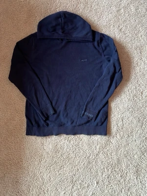 Mörkblå hoodie från pepe jeans - Mörkblå hoodie från pepe jeans med diskret broderad logga på bröstet. Tillverkad i mjukt bomullsmaterial, har ribbade muddar vid ärmslut och nederkant samt klassisk huva. Perfekt för en avslappnad och stilren look.