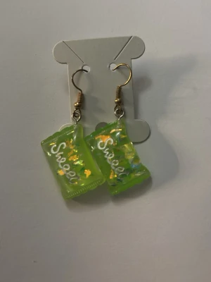 Gröna örhängen med godisdesign - Unika örhängen i form av gröna godispåsar med texten 'Sweet' och små färgglada detaljer inuti. Hängande modell med guldfärgade krokar. Perfekta för dig som gillar lekfulla accessoarer och vill sticka ut.
