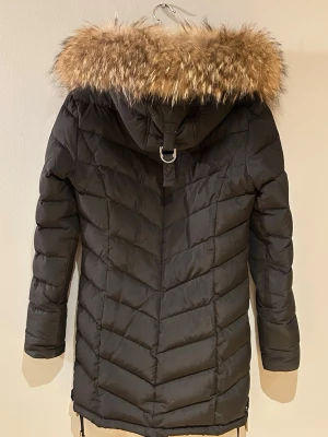 Svart lång dunjacka med äkta päls  - Hej säljer en svart dunjacka i längre modell  med huva och äkta päls, från Chelsea i strl 34. Jackan har dragkedja och tryckknappar framtill, två fickor med dragkedja och justerbar nedertill.