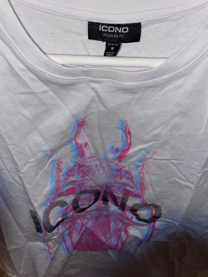 Vit ICONO t-shirt med tryck - Säljer en vit t-shirt från ICONO i regular fit med ett stort, färgstarkt tryck i rosa och blått på framsidan. T-shirten har rund hals och korta ärmar. Perfekt för dig som gillar grafiska motiv och vill sticka ut lite extra. Aldrig använd