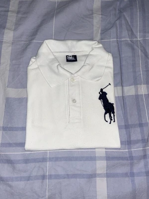 Vit pikétröja från Polo Ralph Lauren - Klassisk vit pikétröja från Polo Ralph Lauren med stor mörkblå broderad logga på bröstet. Tröjan har krage, knappar framtill och är kortärmad. Perfekt för en stilren och sportig look.