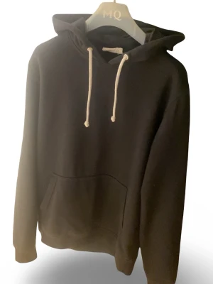 Svart hoodie från Pier One - Klassisk svart hoodie från Pier One med vita snören och stor känguruficka framtill. Mjuk och skön bomullskänsla, perfekt för en avslappnad stil. Huva och ribbade muddar vid ärmslut och nederkant.