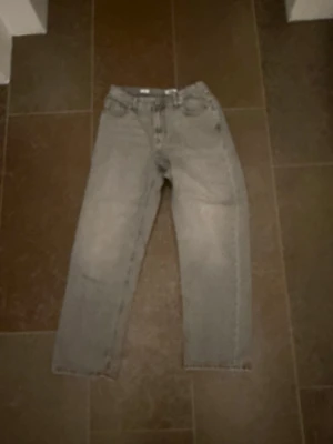 Grå raka jeans i vintage-stil - Säljer ett par grå jeans med rak passform och klassisk femficksdesign. Jeansen har en ljus tvätt och hög midja, perfekta för en avslappnad och trendig look. Materialet är denim i bomull. Passar dig som gillar retrovibbar och enkel stil.