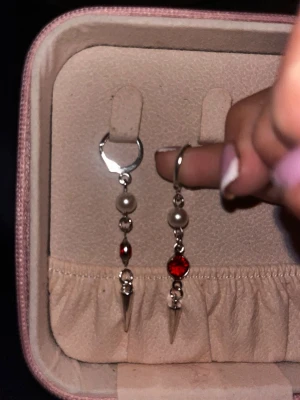 Silvriga örhängen med pärlor och röd sten - Unika silvriga örhängen med hängande detaljer, vita pärlor och en röd glittrande sten. Örhängena har även en liten metallspets längst ner för en edgy look. Perfekta för dig som vill sticka ut med accessoarerna.