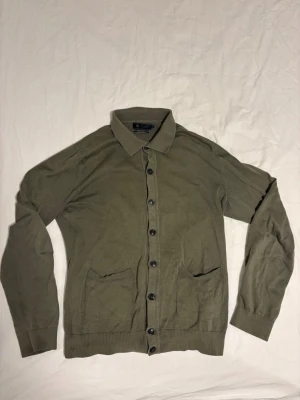 Riley cardigan - Stilren olivgrön kofta med knappar framtill, två fickor och klassisk krage. Långärmad modell i mjukt, tunt stickat material som passar perfekt till lager på lager. Ribbstickade muddar vid ärmslut och nederkant för en snygg passform. Tillverkas inte längre!