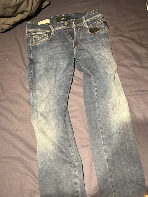 Replay Anbass jeans blå, storlek 29/32 - Snygga Replay Anbass jeans i klassisk blå tvätt med coola slitningar och kontrastsömmar. Modellen har fem fickor, svarta läderdetaljer och en slim passform. Materialet är stretchigt jeans, vilket gör dem riktigt sköna att ha på sig.